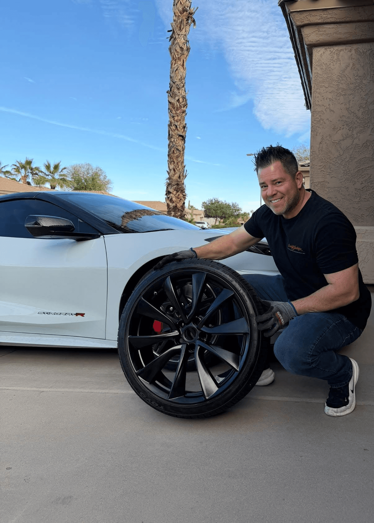 Larri customer kneeling beside a refinished black wheel and tire next to a white sports car